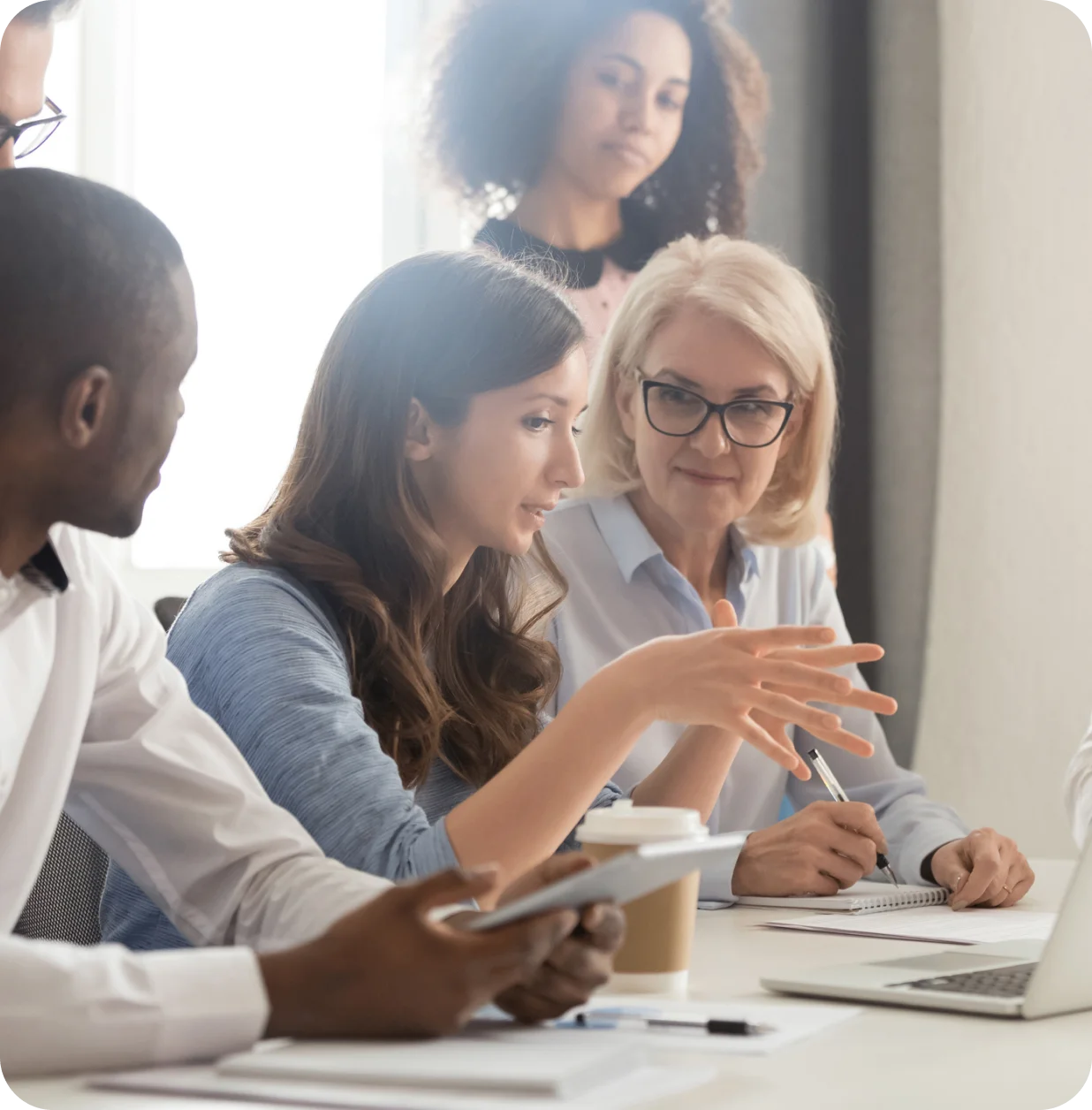 h2 1 A group of four people is engaged in a discussion around a table with a laptop, tablet, notebook, and coffee cup. One person is speaking while the others are listening attentively.